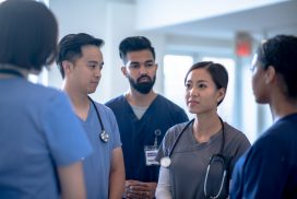 A small group of 5 medical professionals meet casually in the hallway of a hospital to discuss a patient case.  They are each dressed professionally in medical scrubs and are paying close attention as the one nurse brings forth her concerns to the group.